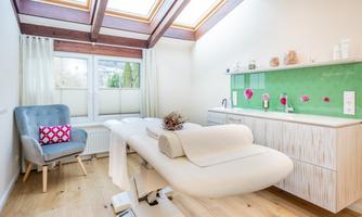 A bright treatment room with a massage table and large windows. Decorative elements and care products are arranged on a modern shelf along the wall.