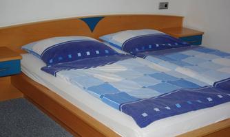 A comfortable double bed with blue and white bedding. The background features a simple and modern bedroom environment.