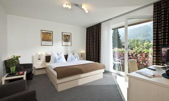 A modern hotel room with a double bed, comfortable armchairs, and a desk. The balcony offers a view of the green landscape.