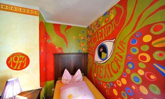 A colorfully designed room with a striking mural in the colors red, green, and yellow. The room has a bed with a pink pillow and a purple lamp.