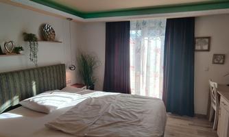 A cozy bedroom with a large bed and wooden details. Bright curtains let in daylight, and plants create a relaxing atmosphere.