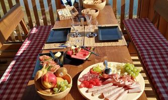 A set table with various appetizing dishes, including fruit and sausage platters. In the background, a calm lake and a beautiful view of the mountains can be seen.