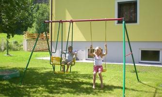 A swing in the garden with two children. In the background, there is a light yellow house and a green meadow.