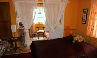 A cozy room with orange walls and a large bed. Next to the window, there is a chair and a small table.