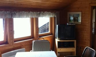 A cozy room with wooden walls and large windows. A table with a checkered tablecloth and a TV are also visible.
