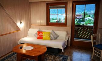 A cozy room with a white sofa, a wooden table, and colorful cushions. Through the window, one looks out at a green landscape and mountains.