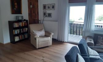 A cozy living room with a comfortable armchair and a bookshelf. Large windows let in plenty of daylight and provide access to a balcony.