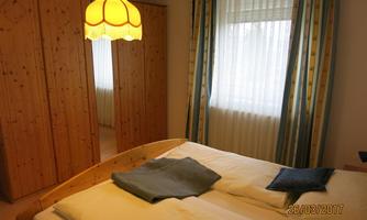 A cozy bedroom with a double bed and a wooden wardrobe. The window lets in a lot of light and a lamp hangs from the ceiling.
