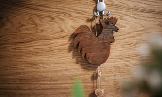 A wooden rooster hangs on a hook in front of a wooden surface. Below it is a small heart pendant.