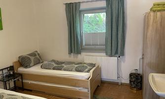 A cozy bedroom with two beds and a window that lets in light. The walls are bright and the windows are decorated with checkered curtains.