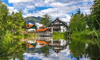 A picturesque house by the water with surrounding greenery. The clouds are calmly reflected in the pond.