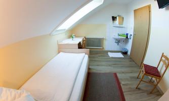 A simple room with a single bed and a small table. The walls are painted in light colors, and there is a window that lets in a lot of light.