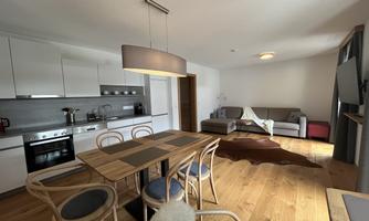 A modern living room with a dining area and an open kitchen. The room is bright and cozy, furnished with wooden flooring and a large window.