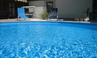 A clear blue pool with sun umbrellas and lounge chairs. In the background, there is a bright house and some plants.