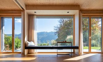 A modern room with large windows offering a view of the green landscape and the mountains. The room has a bright wooden ceiling and a simple arrangement of furniture.