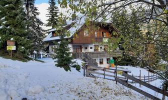 A cozy wooden house is situated in a snowy landscape. Surrounded by fir trees, the scene radiates a tranquil winter atmosphere.