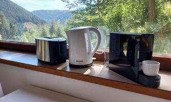 A modern kitchenette with a toaster, a kettle, and a coffee machine. In the background, there is a beautiful view of the mountains and trees.