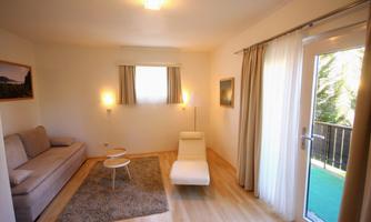 A bright living room with a beige sofa and a modern reclining chair. Large windows provide plenty of daylight and a friendly atmosphere.