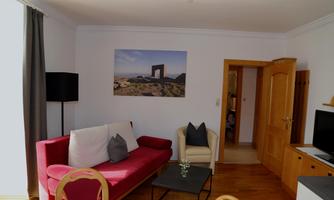 A cozy living space with a red sofa and a comfortable armchair. A picture hangs on the wall and the door leads to another room.