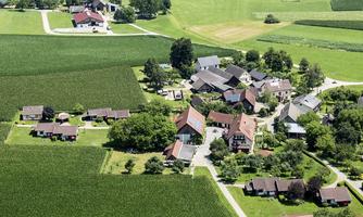 An idyllic landscape with a small village surrounded by green fields. The houses are scattered and surrounded by trees.