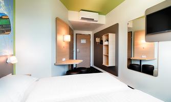 A modern hotel room with two beds, a small table, and a television on the wall. The walls are designed in bright colors, and there is enough light.