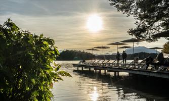 A tranquil lake with a wooden pier and sun loungers. The sun shines brightly over the horizon and reflects in the water.