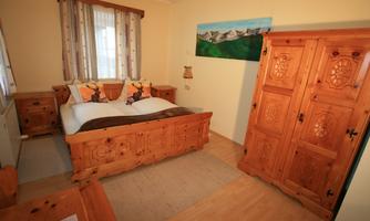 A cozy bedroom with a large wooden bed and rustic furniture. A landscape painting hangs on the wall and lots of daylight streams through the window.