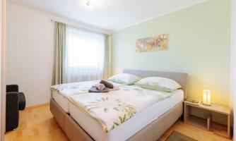 A bright bedroom with a double bed and fresh, green walls. The room is cozily furnished and features large windows that provide plenty of daylight.
