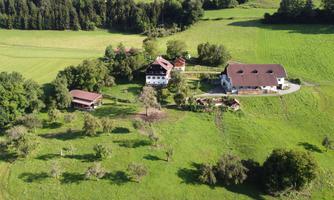 A rural scene with a farmhouse, surrounded by green meadows and trees. In the background, gentle hills and forests can be seen.
