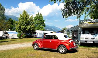 A red car is parked at a campsite, surrounded by motorhomes and trees. The sky is blue with a few clouds.