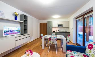 A modern living room with a bright decor and a cozy dining area. The room has large windows and is well-lit.
