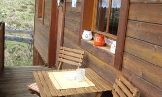 A cozy balcony with a small wooden table and two chairs. The walls are made of wood, and there is a window with curtains.