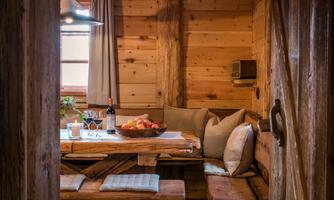 A cozy dining area in a wooden cabin with a wooden table and benches. On the table, there is a fruit bowl, a bottle of wine, and some glasses.