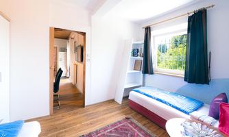 A bright and inviting room with wooden floors and large windows. There is a bed, shelves, and an armchair, as well as a view outside.