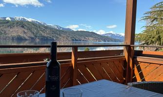 A table with a bottle of wine and glasses on a terrace overlooking the lake and the mountains. The sky is clear and blue.