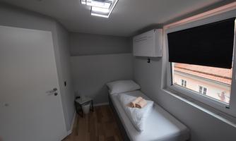 A simple bedroom with a single bed and a small nightstand. The window offers a view outside, and the walls are painted light.