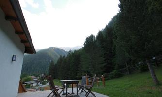 A cozy outdoor area with a table and chairs, surrounded by tall trees and mountains in the background. The sky is slightly cloudy, creating a relaxed atmosphere.