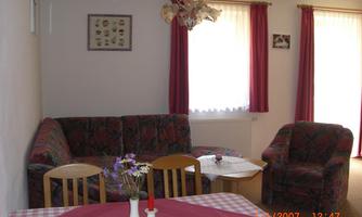 A living room with a cozy sofa and an armchair. A table with a tablecloth and flowers is in the foreground.