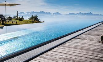 An infinity pool with sparkling water and a magnificent view of the mountains. Nearby, there are deck chairs and a sunshade.