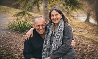 A happy couple is sitting outdoors and smiling at the camera. In the background, trees and a green meadow are visible.
