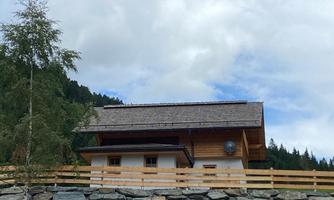 A modern wooden house with a stone wall and a fenced garden. In the background, green hills and forests are visible.