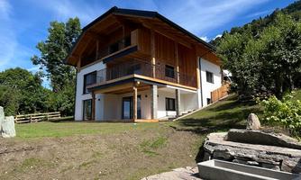 A modern house with wooden cladding and a large balcony. It is located in a green environment with trees and a clear blue sky.