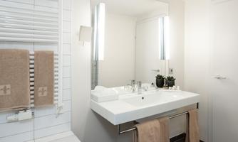 A modern bathroom with a large mirror and a white sink. Towels hang nearby, and there are some plants on the shelf.