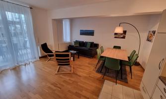 A modern living room with wooden floors and large windows. There is a seating area with chairs and a table, as well as a cozy area with a couch.