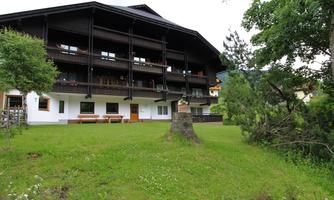 A large, traditional wooden house with several balconies is located in a green environment. The garden is well-maintained and surrounded by trees.