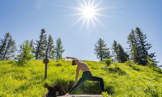 A person is doing yoga outdoors on a meadow. In the background, there are trees and the sun is shining brightly in the sky.