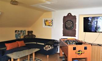 A cozy game room with a blue sofa, a table, and a foosball table. Artworks hang on the wall, and a TV shows a forest.