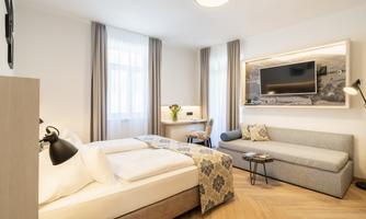 A modern hotel room with two beds, a cozy sofa, and a desk. Large windows allow plenty of daylight in.
