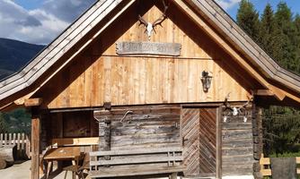 A rustic wooden house with a peaked roof shape in a natural environment. In the background, there are trees and a blue sky.