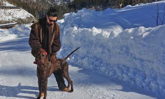 A man stands on a snow-covered path and pets his dog. In the background, snow-covered mountains are visible.